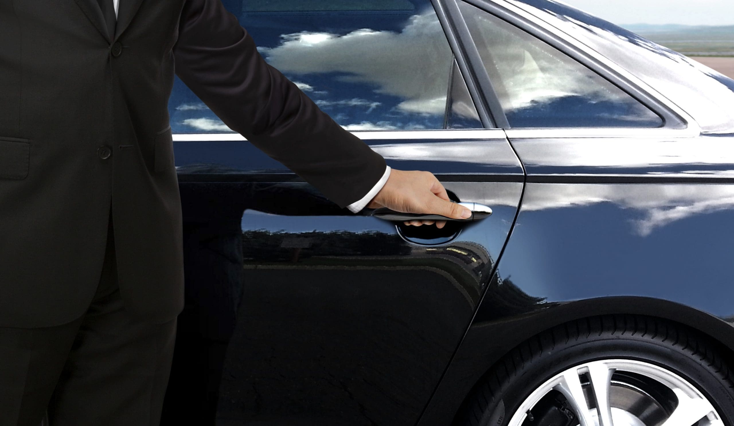 A person in a black suit opens the rear door of a shiny black car. The car reflects clouds and the sky, with part of the driver's side visible. The scene suggests a professional or formal setting.