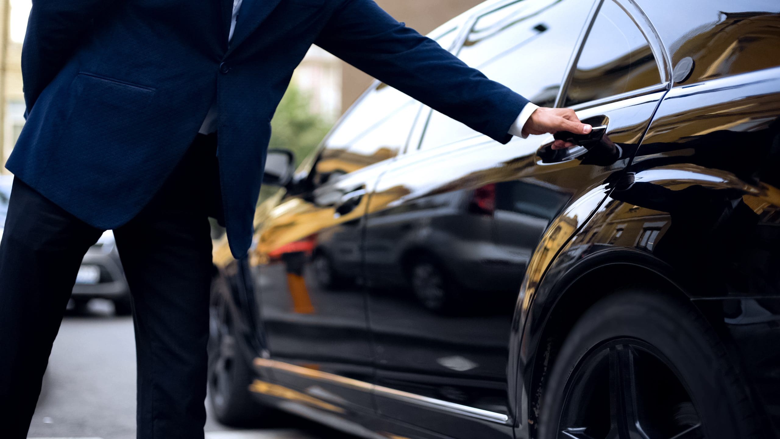 A person in a blue suit opens the rear door of a shiny black car parked on a street with blurred buildings and other cars in the background. The car reflects its surroundings.