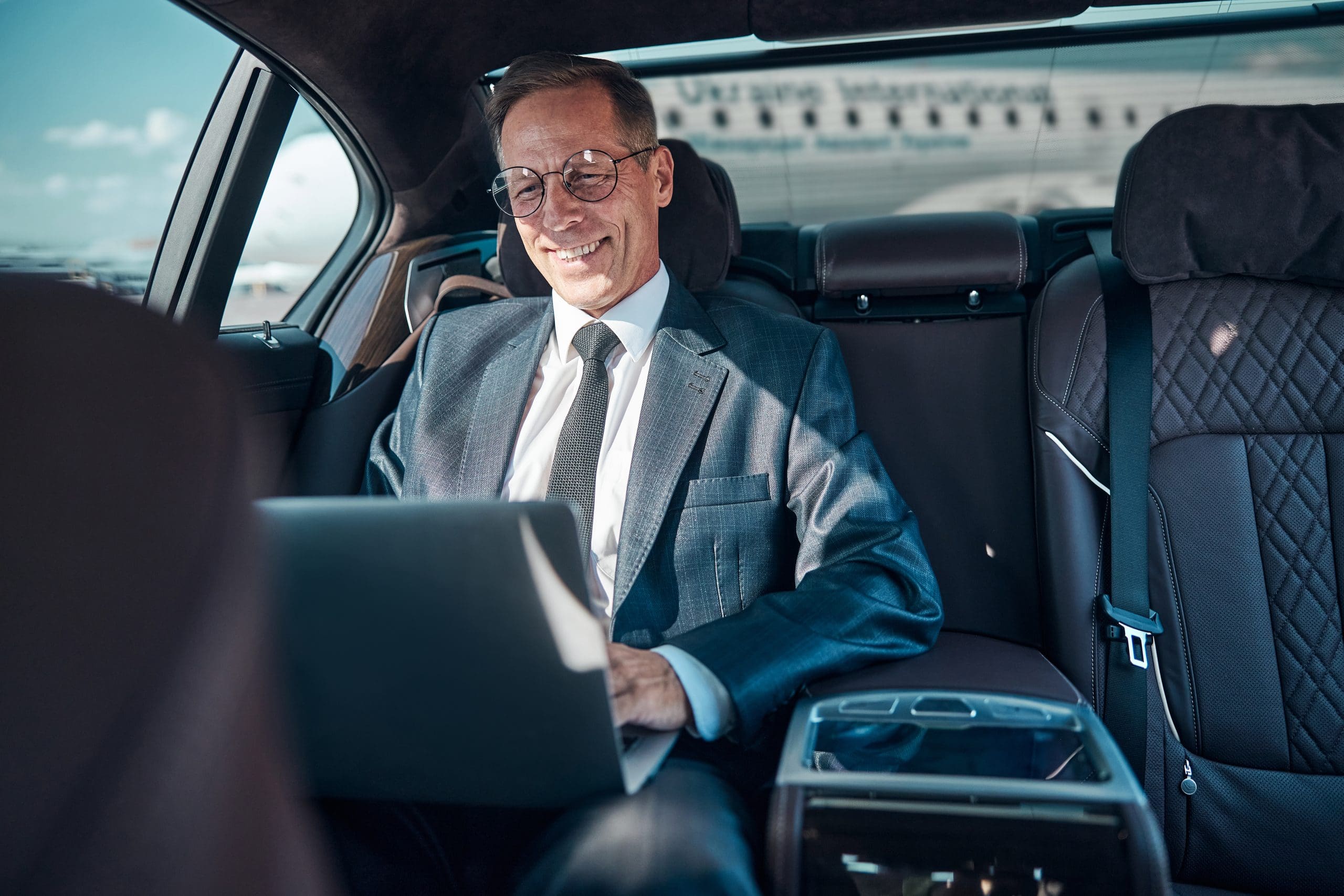 A smiling man in a suit works on a laptop in the backseat of a car. Sunlight streams through the window, illuminating his face. Seats are upholstered in dark leather, and there's a cityscape visible outside.