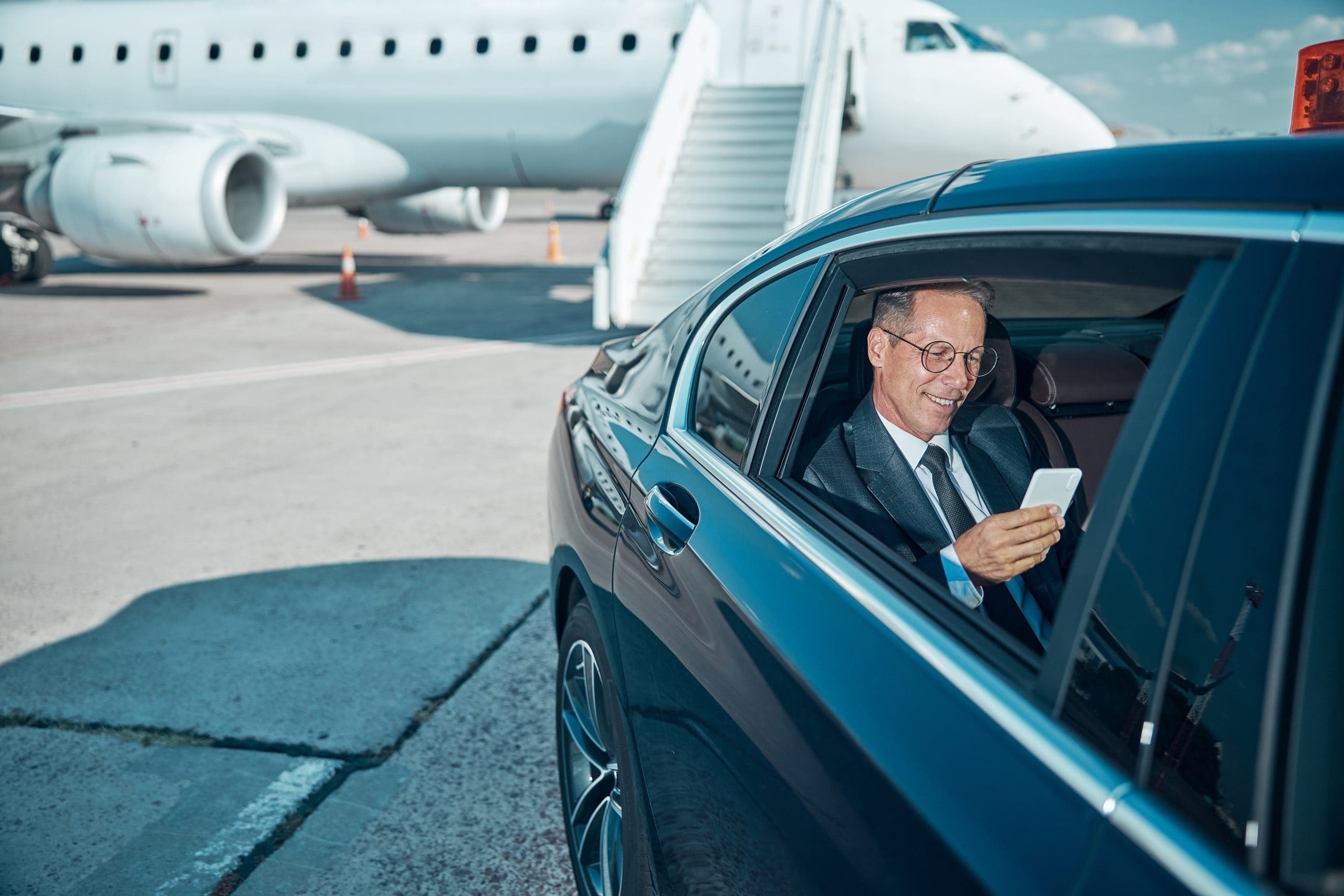 A man in a suit and glasses sits in the back seat of a car, smiling while looking at his smartphone. The car is parked on an airport tarmac near a stairway leading to a parked airplane.