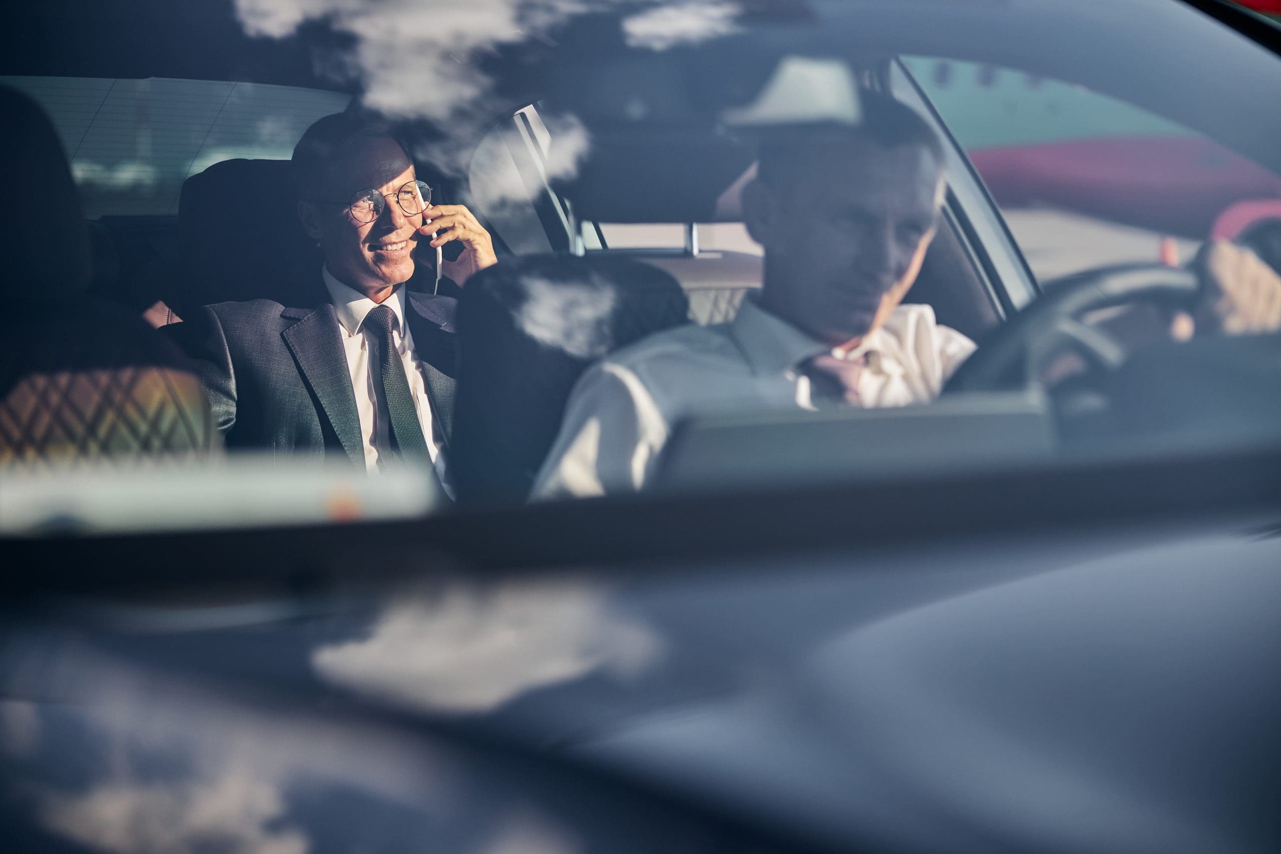 Two men are inside a car. The man in the backseat is smiling and talking on a phone, wearing a suit and glasses. The driver is focused on the road, wearing a white shirt. Reflections and shadows are visible on the car's windshield.