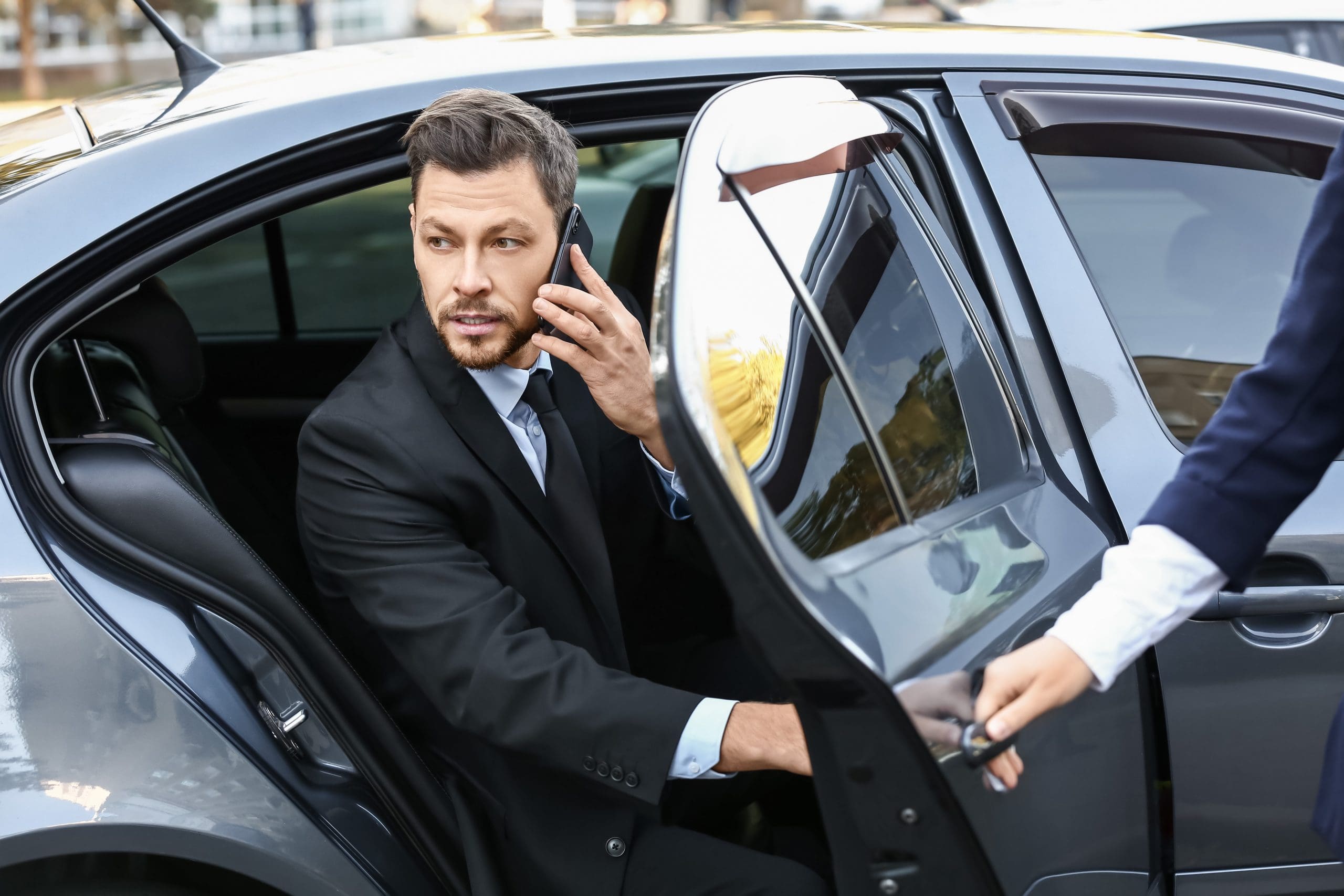 A man in a suit is sitting in the back seat of a car, holding a phone to his ear. The car door is open, and another person, partly visible, is holding the door. The scene appears to be in an urban setting.