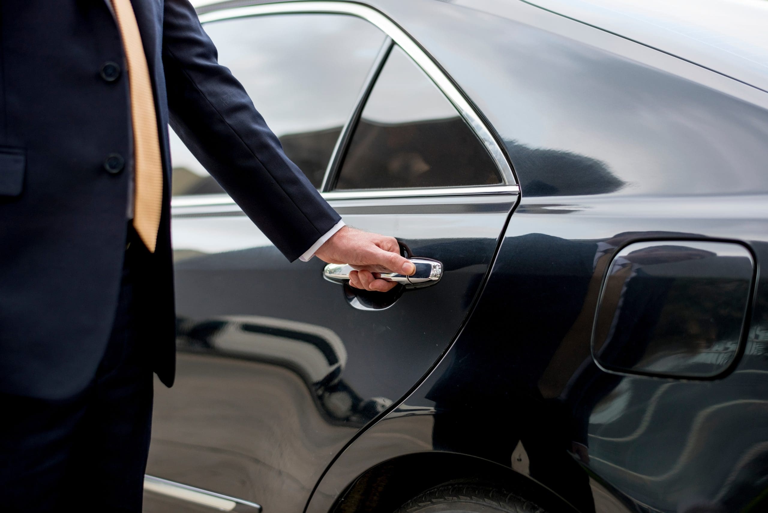 A person in a suit opens a black car door, holding the handle. The side and part of the rear window of the car are visible, with a reflection of the surroundings in the shiny vehicle surface.