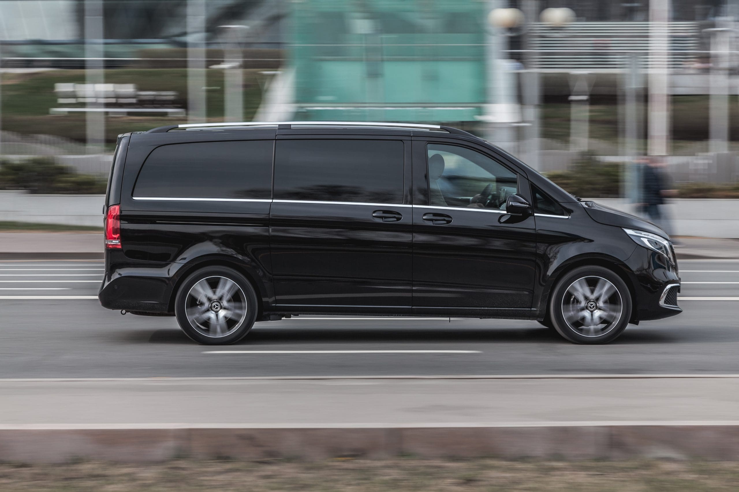 A black minivan is driving on a city street. Its sleek design is visible against a blurred urban background, suggesting motion. The vehicle features tinted windows and silver wheels.