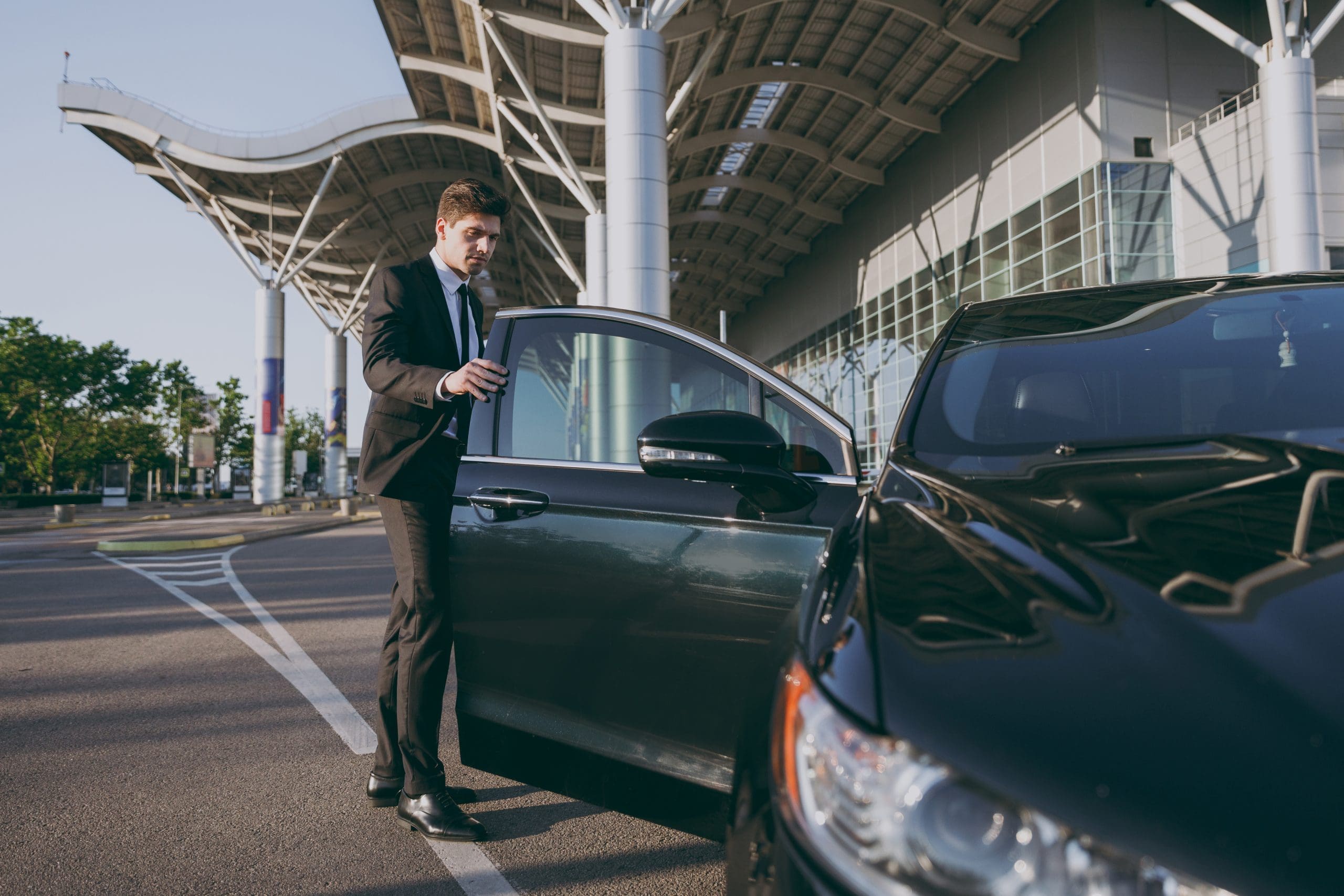 A man in a suit stands next to an open car door in a modern parking area. He appears to be getting into or out of the black car. The background features a contemporary building with large glass windows.