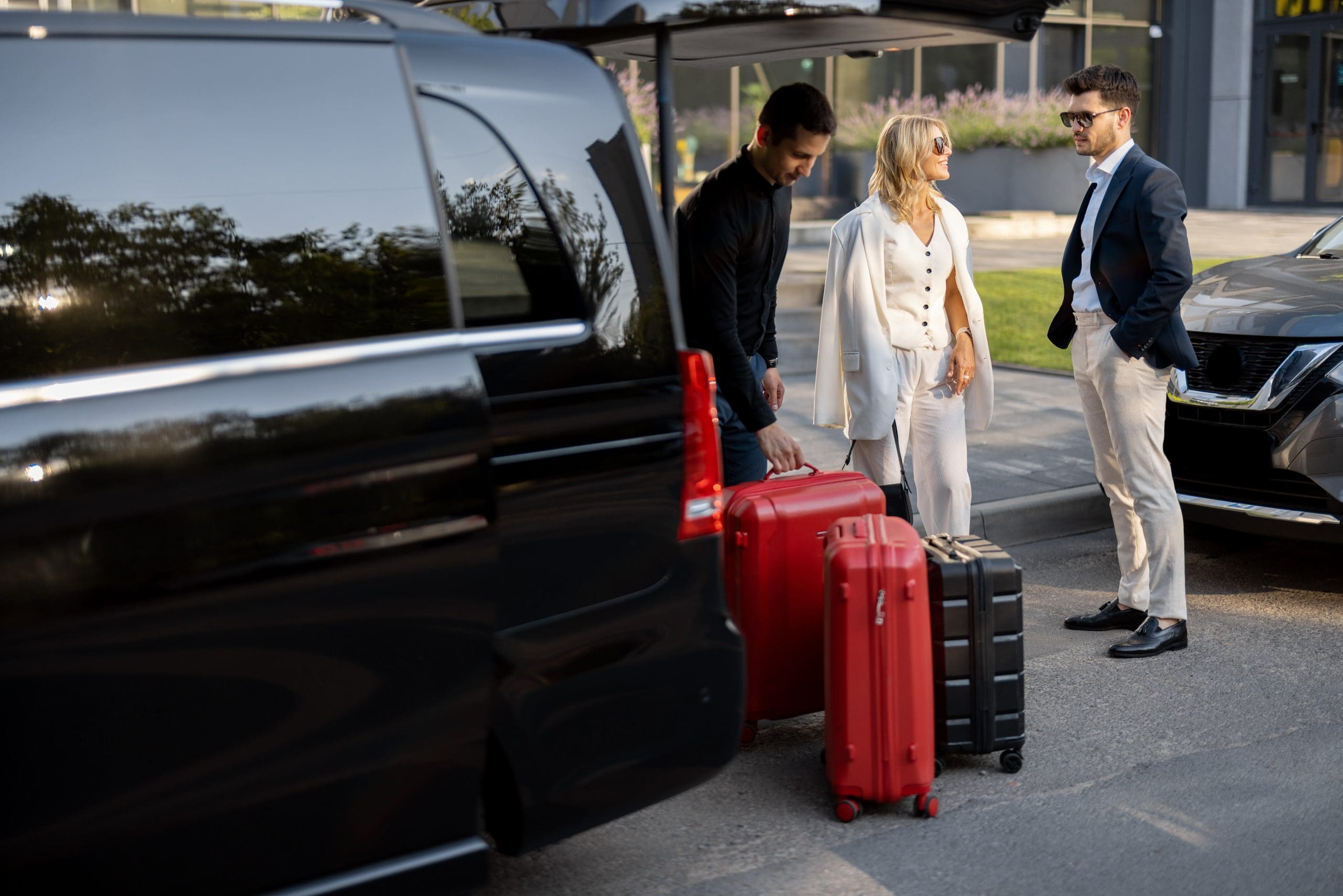 Two men and a woman stand near a black vehicle with its trunk open. They are handling red and black suitcases, suggesting they are getting ready to travel. All are dressed in casual to smart-casual attire.