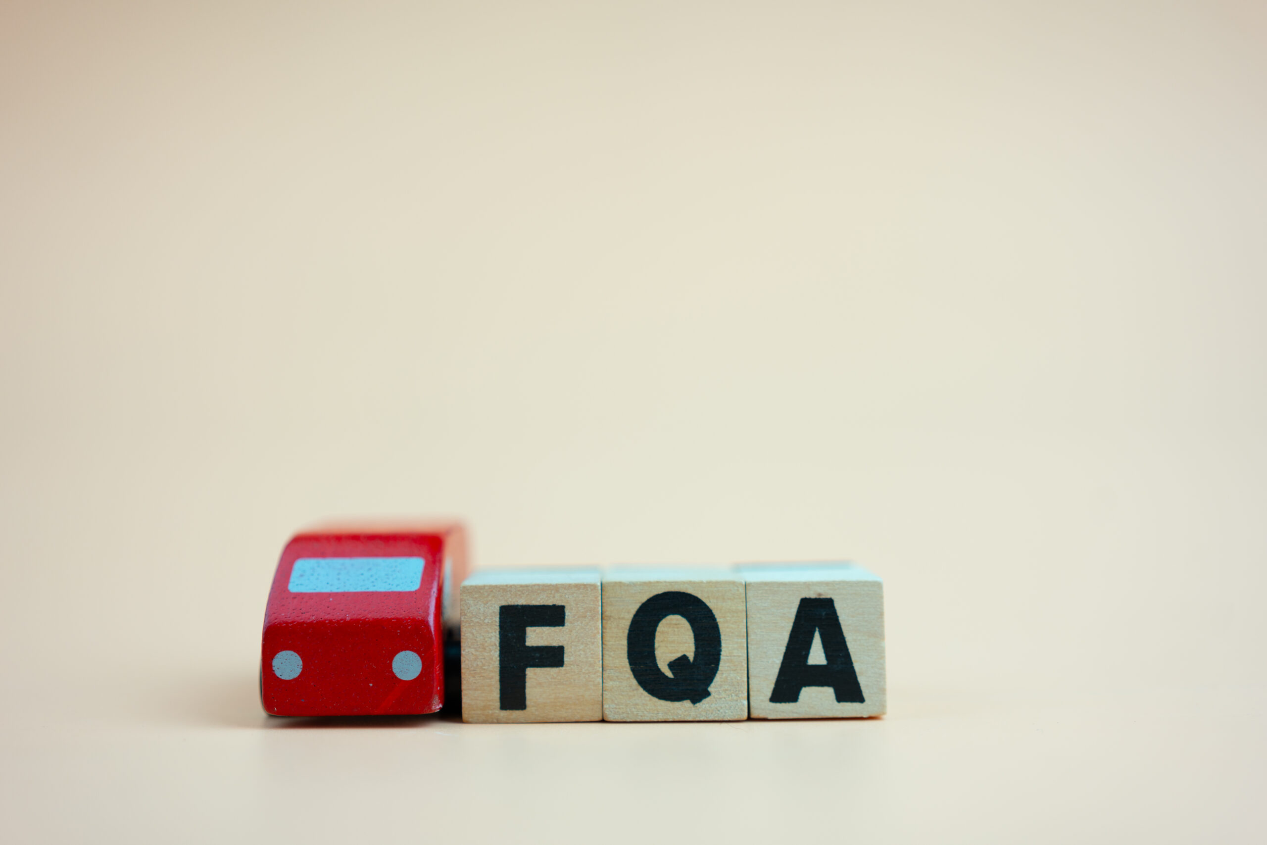 Three wooden blocks with the letters "FQA" are arranged in a row, next to a small red toy car, all set against a plain, light-colored background.