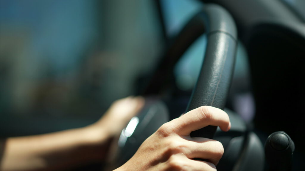 Close-up of a person’s hands gripping a car steering wheel, with a blurred background suggesting the vehicle is in motion. The lighting is bright, highlighting the hands and part of the wheel.