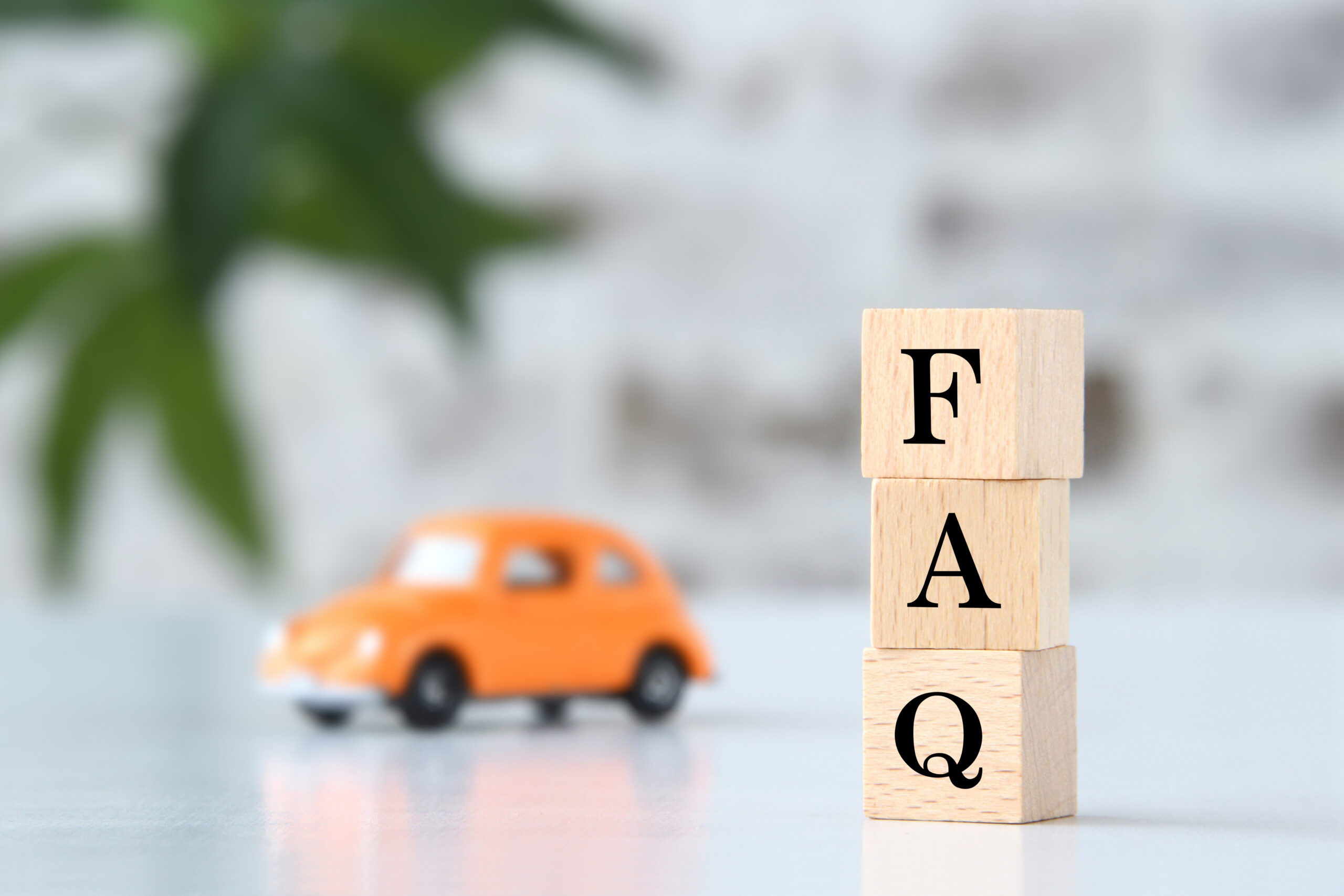 Three wooden blocks stacked vertically spell “FAQ” in black letters, with a blurred orange toy car and green leaves in the background on a white surface.