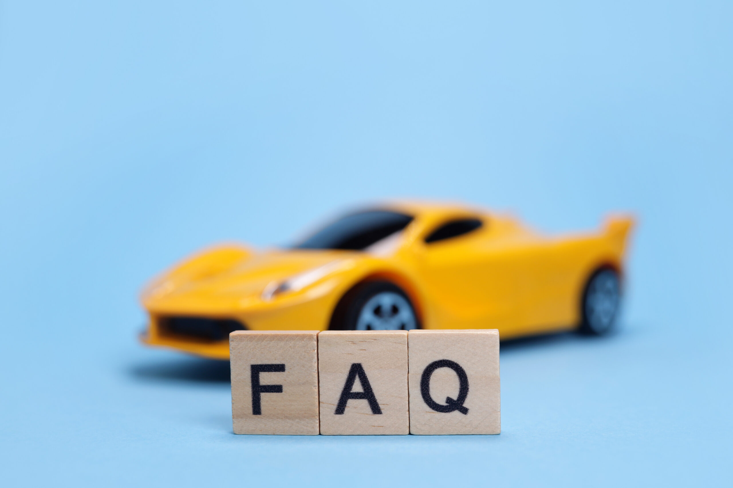 Three wooden blocks with the letters "FAQ" are in focus in the foreground, with a blurred yellow toy sports car in the background against a light blue surface.