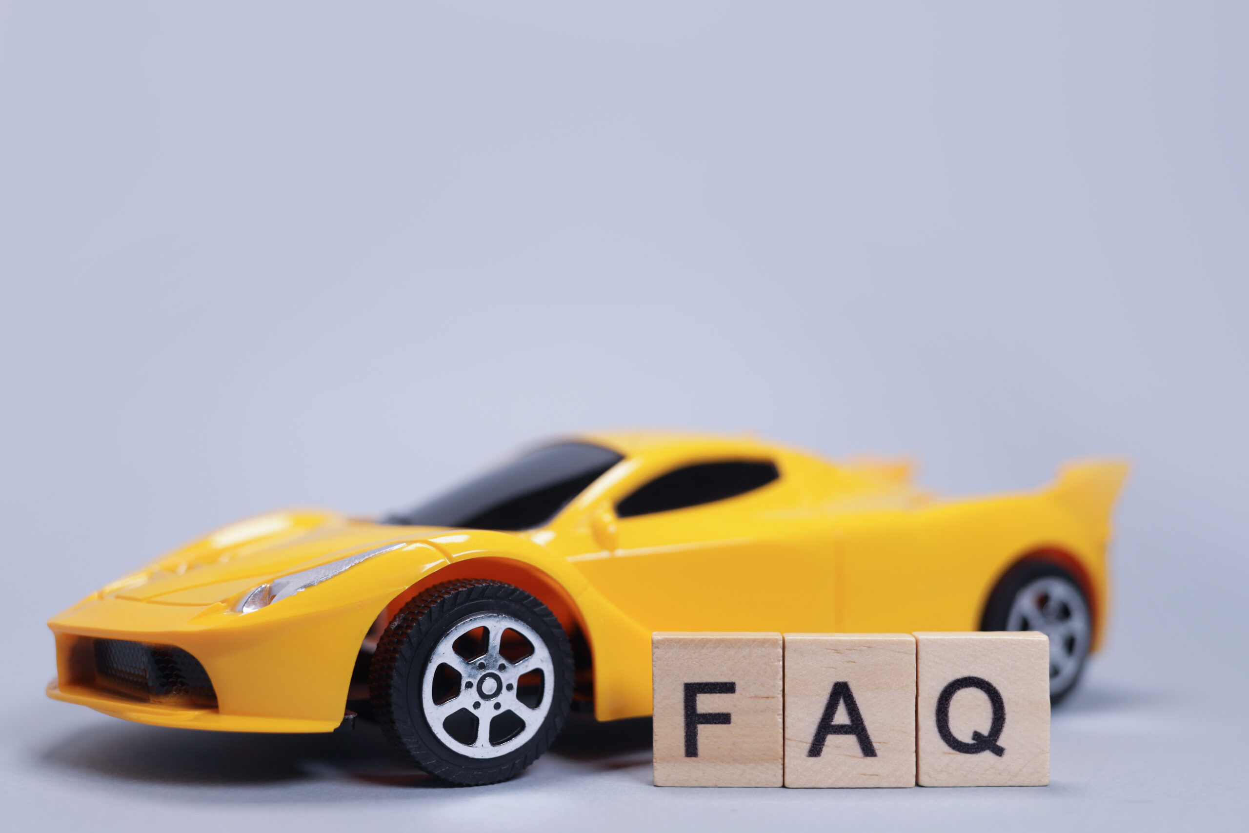A yellow toy sports car is positioned behind three wooden blocks displaying the letters "F", "A", and "Q" against a light gray background.